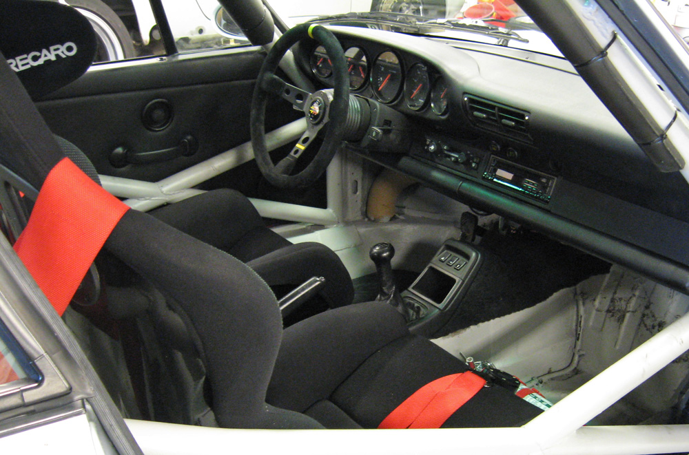 The interior of a Porsche 964 race car showing seats, harnesses and roll cage.