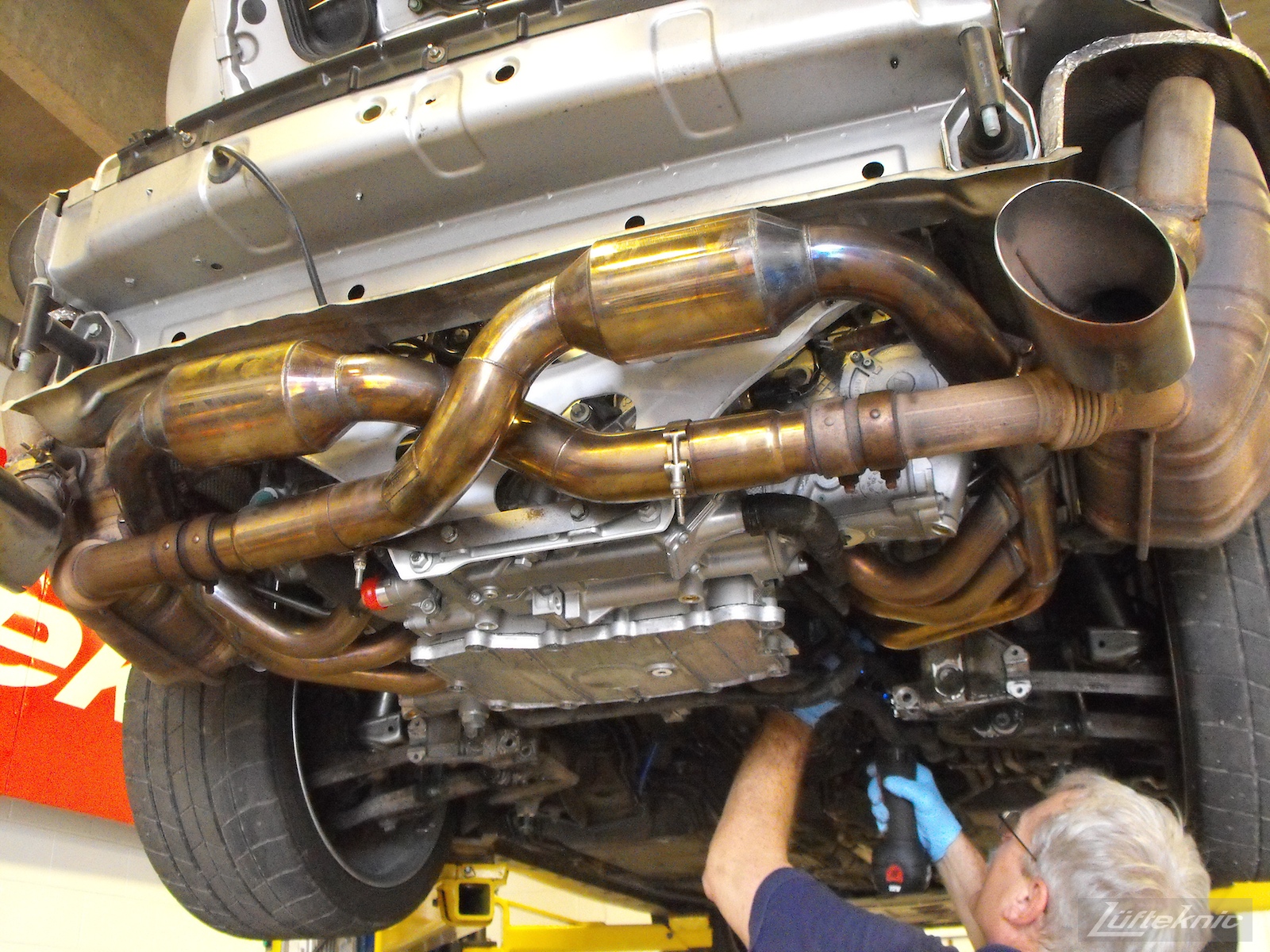 A Porsche 996 Turbo on a lift with the bumper removed showing the sports exhaust and a technician working on another part of the engine.