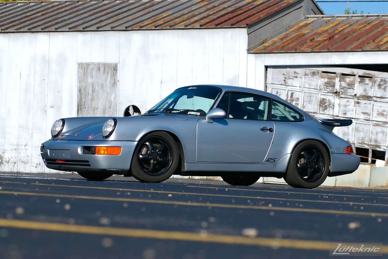 The 964 RS America shown from the front in a parking lot with a dilapidated garage in the background.