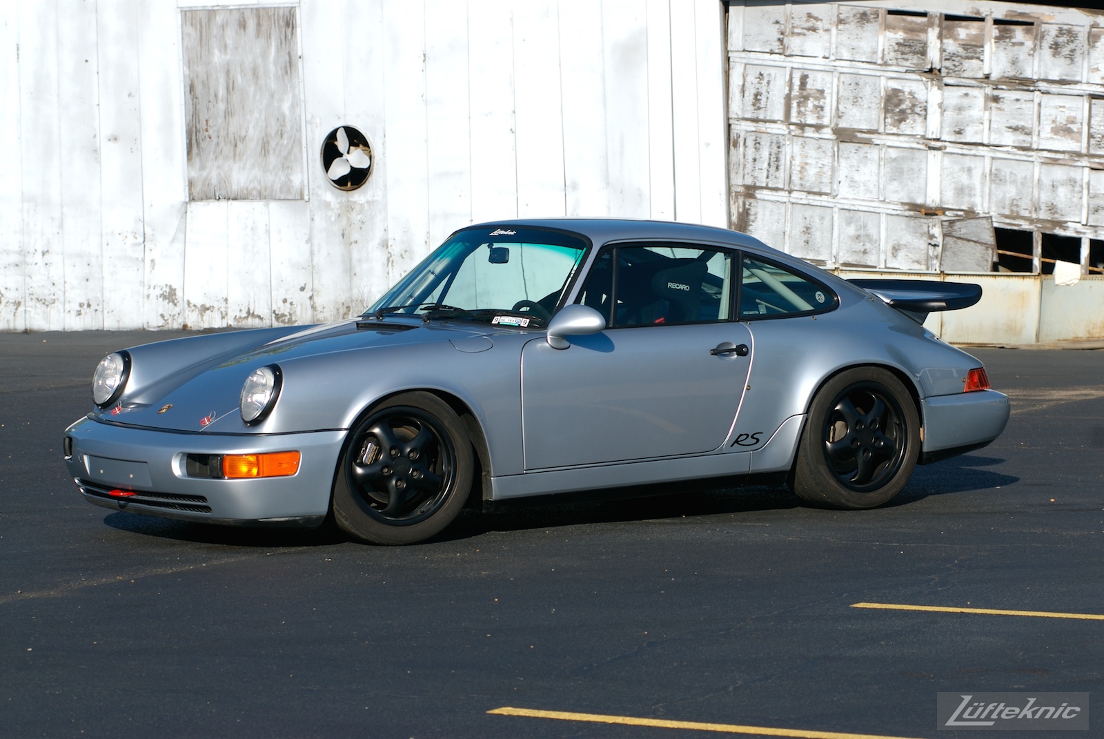 The 964 RS America in a parking lot with a dilapidated garage in the background.