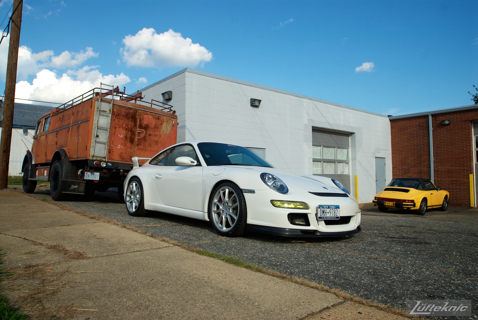 A white Porsche Gt3 poses with the Lufteknic fire truck in the parking lot, with an old yellow 911 Targa in the background.