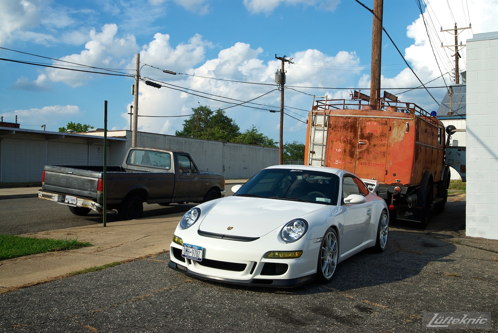 A white Porsche Gt3 poses with the Lufteknic fire truck in the parking lot, with an Ford in the background.
