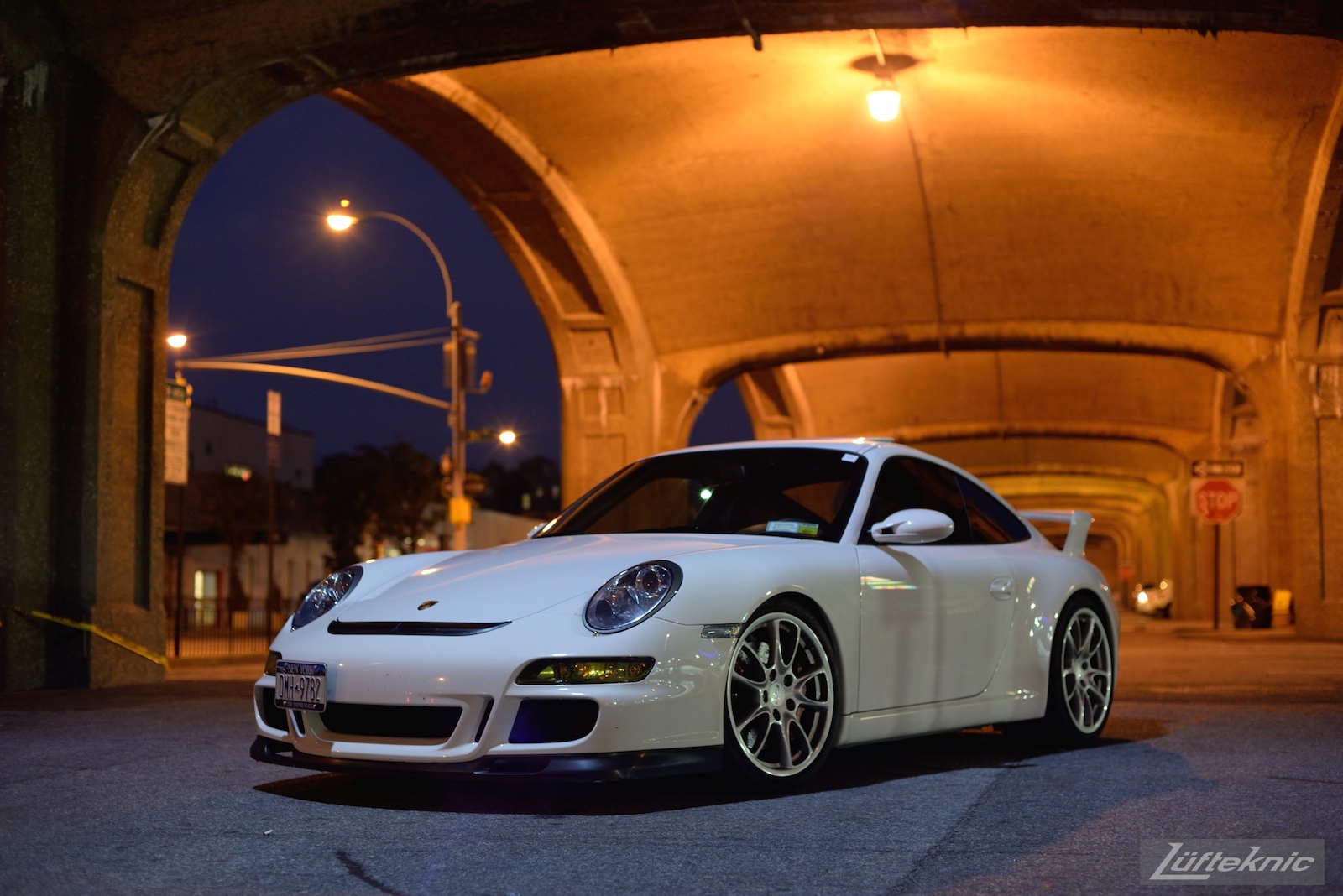 A white 997 GT3 viewed from the front under the 7 train bridge on Queens blvd at night.