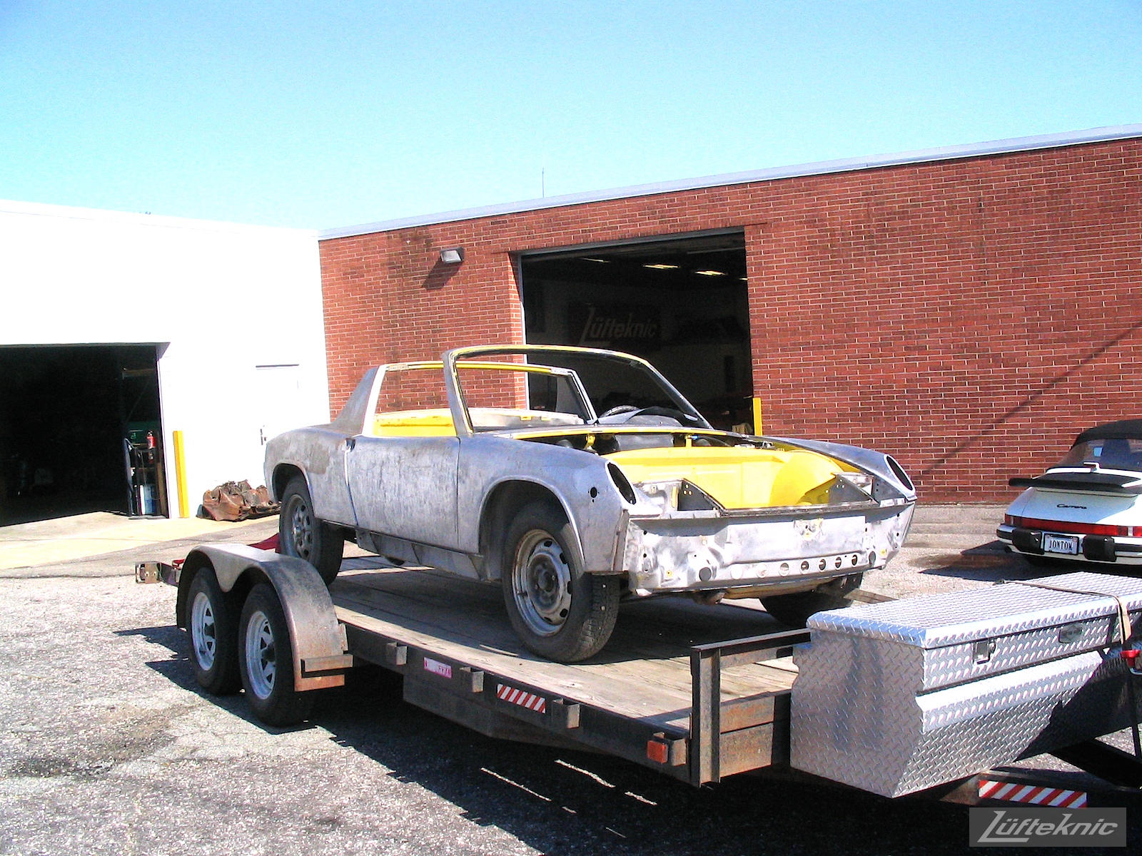 A stripped down yellow Porsche 914 shown mid-restoration on a trailer in the parking lot of Lufteknic.